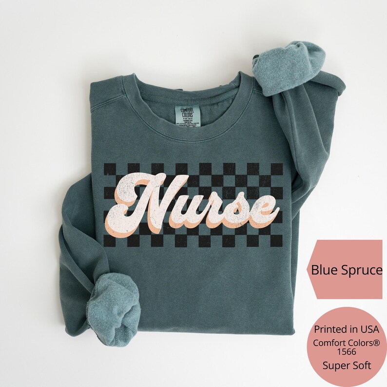 K&ouml;nnte beinhalten: Blau-Fichte-farbenes Sweatshirt mit dem Wort "Nurse" in einem Retro-Schriftzug &uuml;ber einem schwarz-wei&szlig;en Schachbrettmuster. Das Sweatshirt ist in den USA bedruckt und superweich.