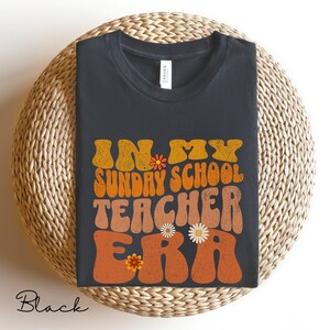 May include: Black t-shirt with a retro-style graphic that says "In My Sunday School Teacher Era" with orange, yellow, and brown colors and floral accents.