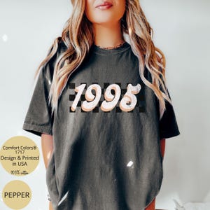 May include: A dark gray t-shirt with a white and black checkered pattern and the year 1995 in a pink and white retro font.