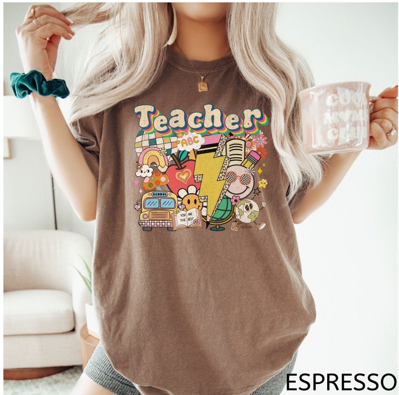 May include: A brown t-shirt with the word "Teacher" in colorful retro letters. The shirt features a graphic design of school-themed illustrations, including a school bus, an apple, a lightning bolt, a globe, and a book.