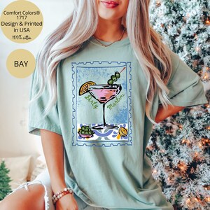 K&ouml;nnte beinhalten: Hellgr&uuml;nes T-Shirt mit einem pinkfarbenen Dirty Martini Cocktail-Motiv in einem blauen Briefmarken-Design. Das Shirt hat auch den Text "Comfort Colors 1717 Design & Printed in USA 100% cotton" und "BAY".
