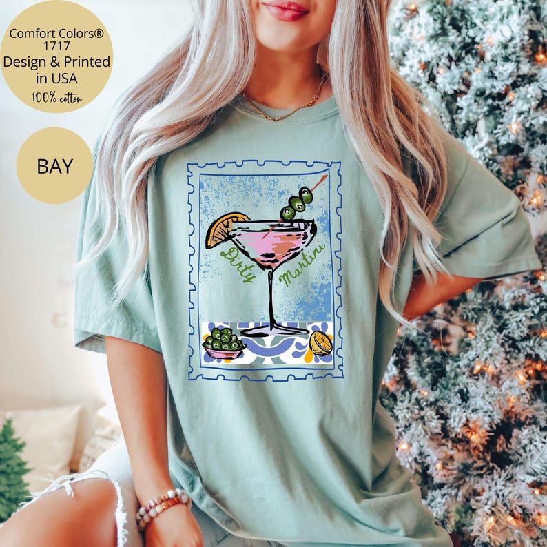 K&ouml;nnte beinhalten: Hellgr&uuml;nes T-Shirt mit einem pinkfarbenen Dirty Martini Cocktail-Motiv in einem blauen Briefmarken-Design. Das Shirt hat auch den Text "Comfort Colors 1717 Design & Printed in USA 100% cotton" und "BAY".