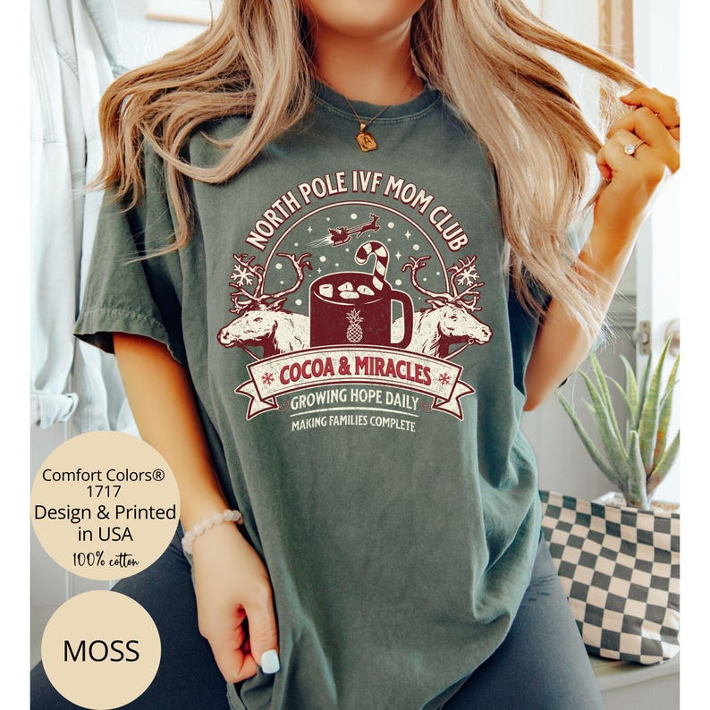 Puede incluir: Una camiseta verde con un dise&ntilde;o navide&ntilde;o que dice "North Pole IVF Mom Club" con una taza de chocolate caliente y renos. El texto debajo del dise&ntilde;o dice "Cocoa & Miracles", "Growing Hope Daily", y "Making Families Complete".