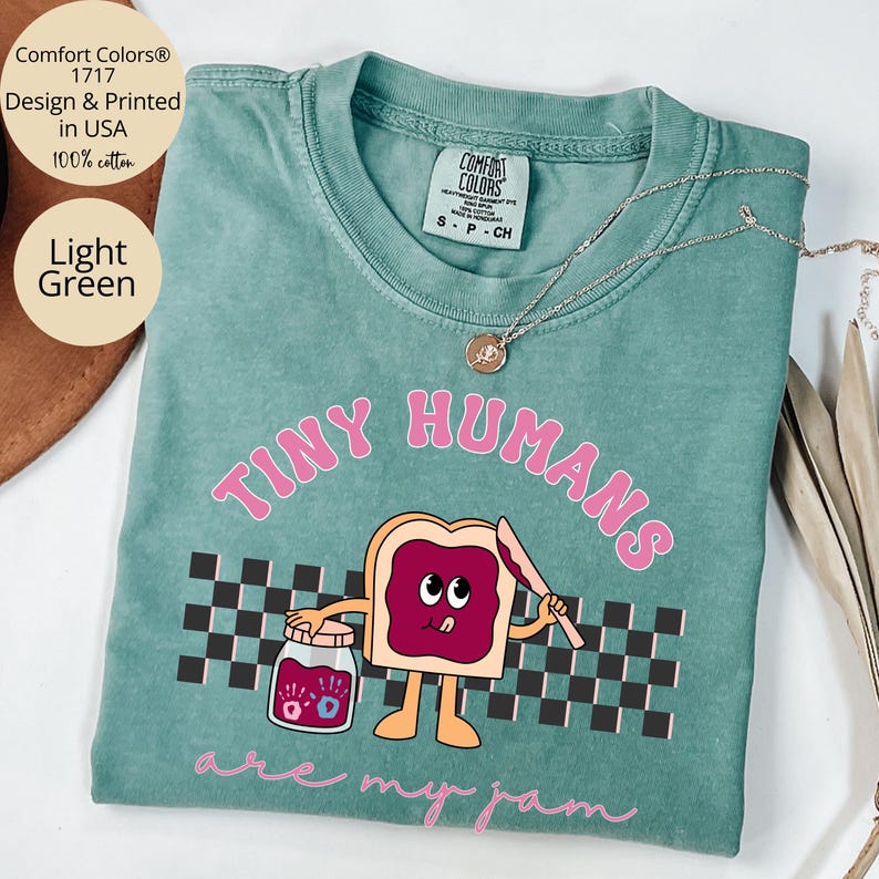 Pu&ograve; includere: T-shirt Comfort Colors verde chiaro con la scritta "TINY HUMANS are my jam" in rosa. La grafica presenta un toast dei cartoni animati con marmellata, un coltello e un barattolo di marmellata. La maglietta &egrave; 100% cotone.