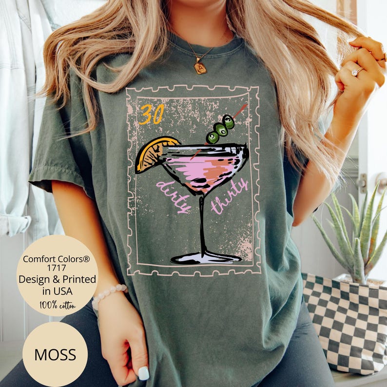 May include: Moss green t-shirt with a graphic of a pink cocktail with olives, a lemon slice, and the words "dirty thirty." The shirt also features the text "Comfort Colors 1717 Design & Printed in USA 100% cotton" and the word "MOSS".