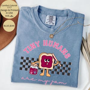 Pu&ograve; includere: T-shirt Comfort Colors blu jeans con la scritta "TINY HUMANS are my jam" in rosa. La grafica presenta un toast a cartoni animati con marmellata, che tiene in mano un coltello e un barattolo di marmellata. La maglietta &egrave; 100% cotone.
