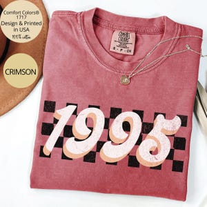 May include: A crimson red t-shirt with a black and white checkered pattern behind the number 1995 in a retro font. The text "Comfort Colors&reg; 1717 Design & Printed in USA 100% cotton" is printed on the top left corner of the shirt.