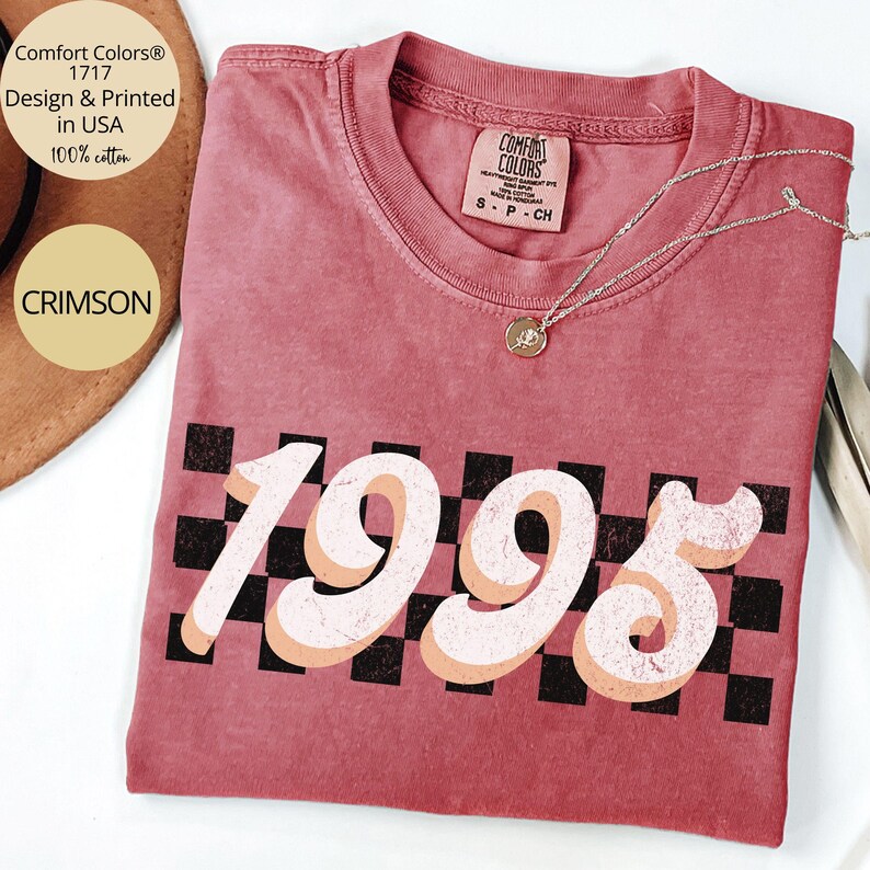 May include: A crimson red t-shirt with a black and white checkered pattern behind the number 1995 in a retro font. The text "Comfort Colors&reg; 1717 Design & Printed in USA 100% cotton" is printed on the top left corner of the shirt.