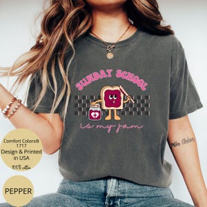 May include: A dark gray t-shirt with the text "SUNDAY SCHOOL is my jam" in pink lettering. The text is accompanied by a cartoon illustration of a slice of bread with a face and a jar of jam with a heart on it.