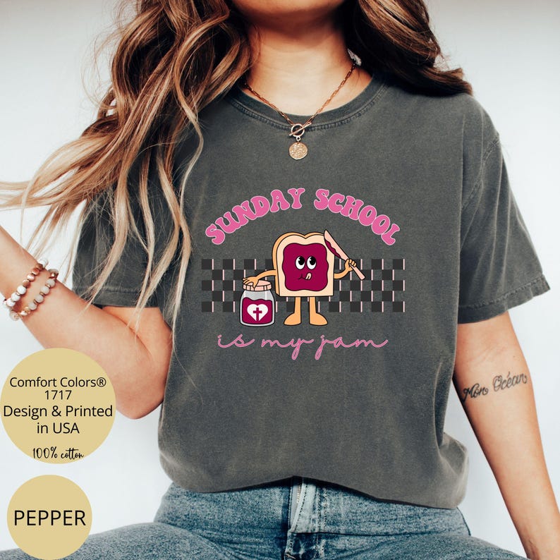May include: A dark gray t-shirt with the text "SUNDAY SCHOOL is my jam" in pink lettering. The text is accompanied by a cartoon illustration of a slice of bread with a face and a jar of jam with a heart on it.