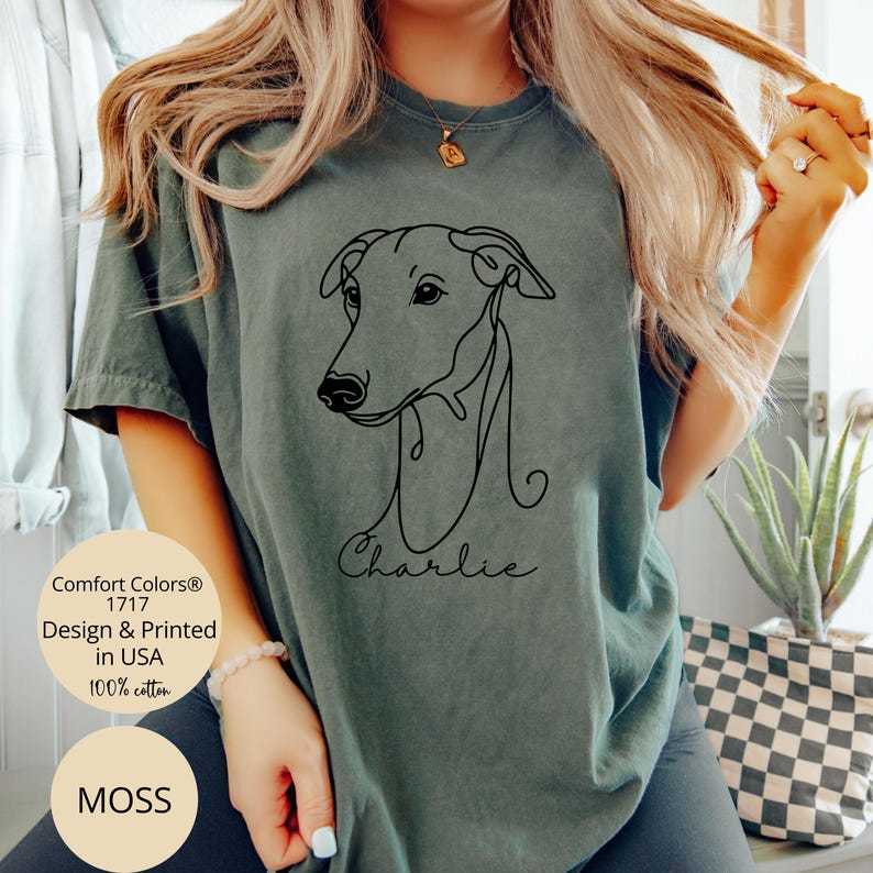 May include: Moss green t-shirt featuring a minimalist black line drawing of a dog's head and the name "Charlie" in script. The shirt is made of 100% cotton and is printed in the USA. The shirt is a Comfort Colors 1717 design.