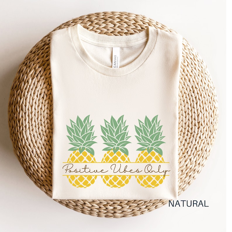 May include: A beige t-shirt featuring a graphic of three yellow pineapples with green tops. The pineapples are arranged in a row with the text "Positive Vibes Only" written in a cursive font below them.