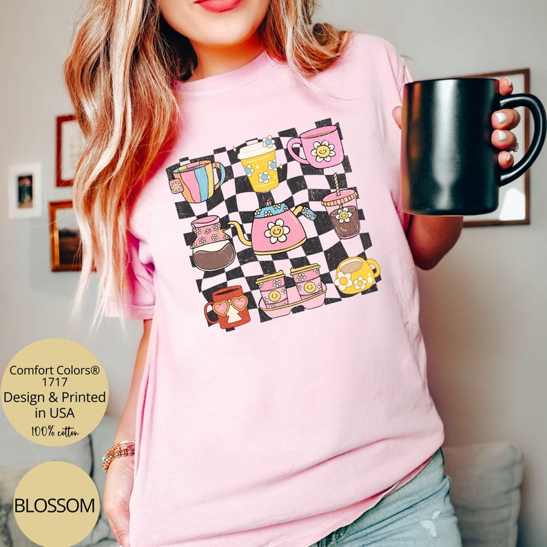 May include: A pink t-shirt with a checkered background and a design of various coffee cups and mugs. The text "Comfort Colors&reg; 1717 Design & Printed in USA 100% cotton" is visible on the bottom left corner of the shirt. The text "BLOSSOM" is visible on the bottom left corner of the shirt.