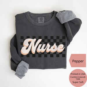 K&ouml;nnte beinhalten: Ein dunkelgraues Sweatshirt mit dem Wort "Nurse" in einem Retro-Schriftzug &uuml;ber einem schwarz-wei&szlig;en Schachbrettmuster. Das Sweatshirt hat ein weiches, fleecegef&uuml;ttertes Innenfutter und die Farbe ist "Pepper". Der Text "Printed in USA Comfort Colors&reg; 1566 Super Soft" ist ebenfalls sichtbar.