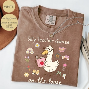 Pode incluir: Uma camiseta marrom com o texto "Silly Teacher Goose on the loose" em branco. A camiseta apresenta um desenho animado de um ganso de &oacute;culos segurando um livro com uma ma&ccedil;&atilde; em cima. H&aacute; outras imagens de desenhos animados ao redor do ganso, incluindo arco-&iacute;ris, estrelas e uma x&iacute;cara de ch&aacute;.