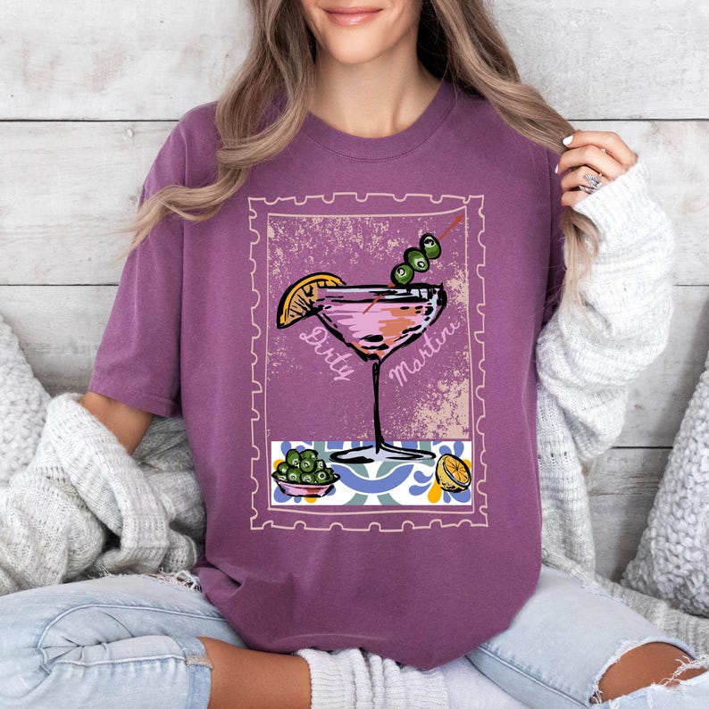 K&ouml;nnte beinhalten: Ein lila T-Shirt mit einer Grafik eines Martini-Cocktails, Oliven und einer Zitronenscheibe. Die Worte "Dirty Martini" sind auf dem Bild aufgedruckt. Das Shirt hat eine einfarbige lila Farbe.