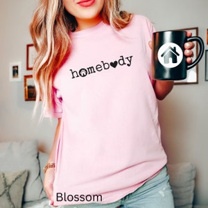 Pode incluir: Uma camiseta rosa com o texto "homebody" em letras pretas com um pequeno &iacute;cone de casa e um cora&ccedil;&atilde;o. A modelo est&aacute; segurando uma caneca preta com um &iacute;cone de casa branco.