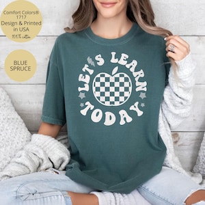 May include: Blue Spruce Comfort Colors t-shirt with the text "LET'S LEARN TODAY" in white. The design includes a checkered apple. The shirt is made of 100% cotton and is designed and printed in the USA.