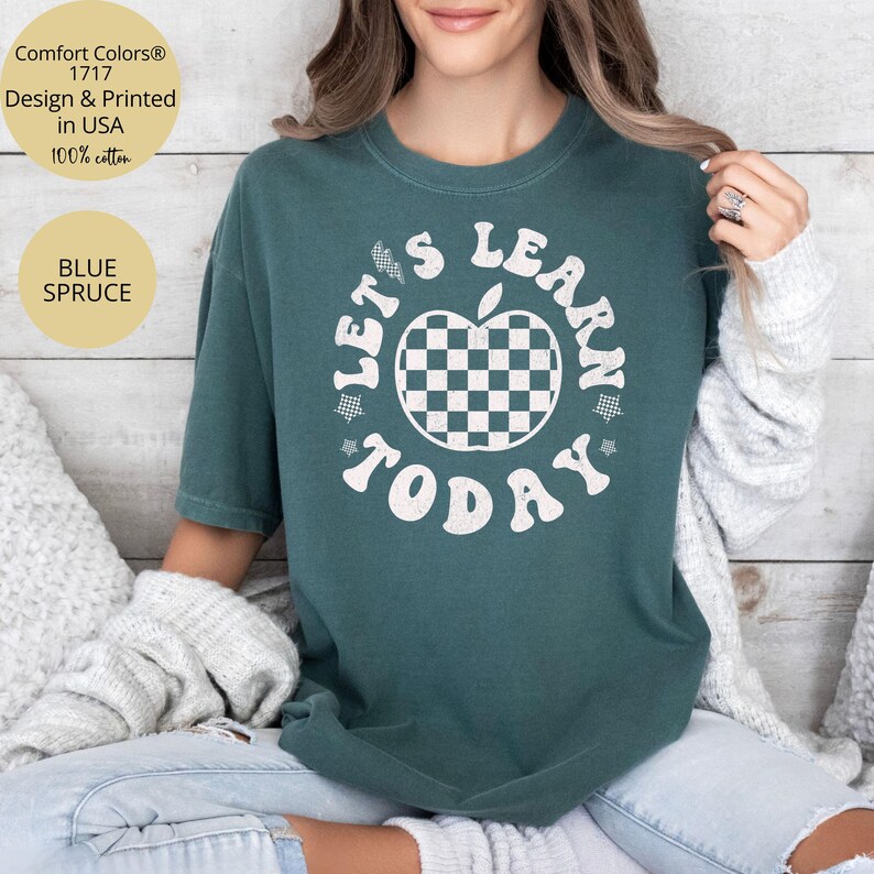 May include: Blue Spruce Comfort Colors t-shirt with the text "LET'S LEARN TODAY" in white. The design includes a checkered apple. The shirt is made of 100% cotton and is designed and printed in the USA.