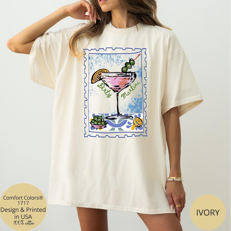K&ouml;nnte beinhalten: Elfenbeinfarbenes T-Shirt mit einer Grafik eines rosa Dirty Martini Cocktails, Oliven und einer Zitronenscheibe, umrahmt von einem blauen Postwertzeichen-Design. Das Shirt zeigt die W&ouml;rter "Dirty Martini" und "Comfort Colors 1717 Design & Printed in USA 100% cotton."