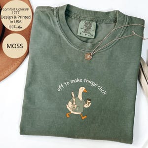 Peut inclure: Un t-shirt en coton vert mousse avec un motif représentant une oie portant une blouse à col en V, tenant un modèle de colonne vertébrale et une tasse de café. Le texte au-dessus de l'oie indique, off to make things click. Un collier doré.