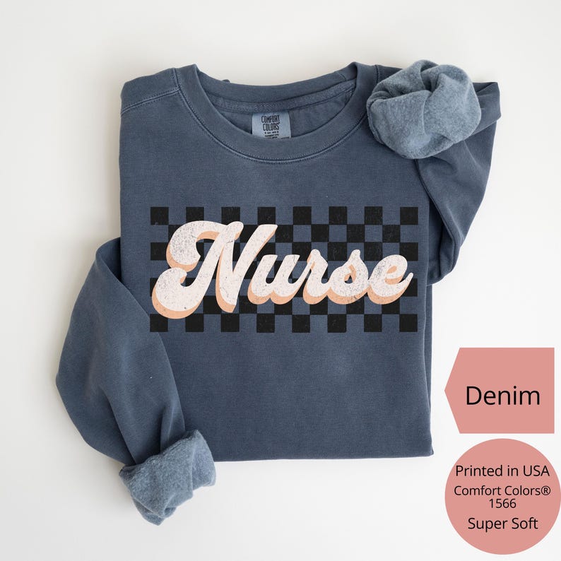 K&ouml;nnte beinhalten: Denimfarbenes Sweatshirt mit dem Wort "Nurse" in einem Retro-Schriftzug &uuml;ber einem schwarz-wei&szlig;en Schachbrettmuster. Das Sweatshirt ist aus einem superweichen Material gefertigt.