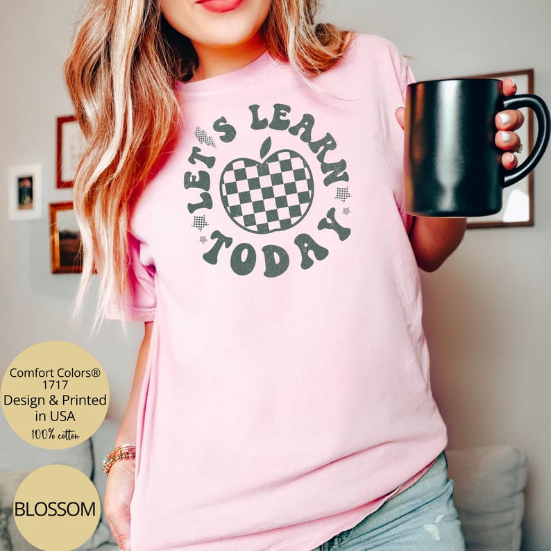 May include: A light pink t-shirt with the text "LET'S LEARN TODAY" surrounding a checkered apple graphic. The shirt is made of 100% cotton and is printed in the USA. A black mug is held in the hand.
