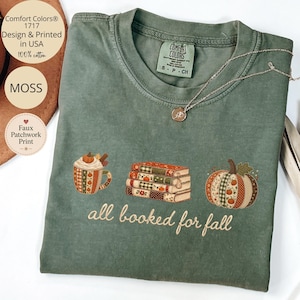 May include: A moss green short-sleeve t-shirt featuring a graphic print of a mug, a stack of books, and a pumpkin, all with a patchwork pattern. The text "all booked for fall" is printed below the graphics in a cursive font. The shirt has a crew neck.