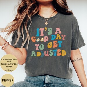Peut inclure: Un t-shirt gris foncé avec un motif coloré qui dit "C'est un bon jour pour s'adapter".