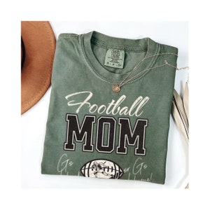 May include: Green t-shirt with the text "Football Mom" in black and white lettering. The shirt also features a vintage-style illustration of a football player.