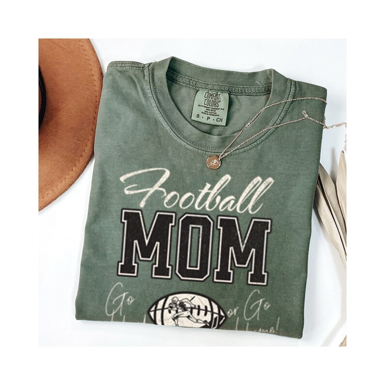May include: Green t-shirt with the text "Football Mom" in black and white lettering. The shirt also features a vintage-style illustration of a football player.