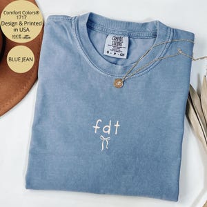 Peut inclure: Un t-shirt bleu jean Comfort Colors avec le texte "fdt" et un motif de n&oelig;ud. Le t-shirt a un col rond et des manches courtes. Un collier en or avec un pendentif est drap&eacute; sur le t-shirt. Le t-shirt est en 100% coton.
