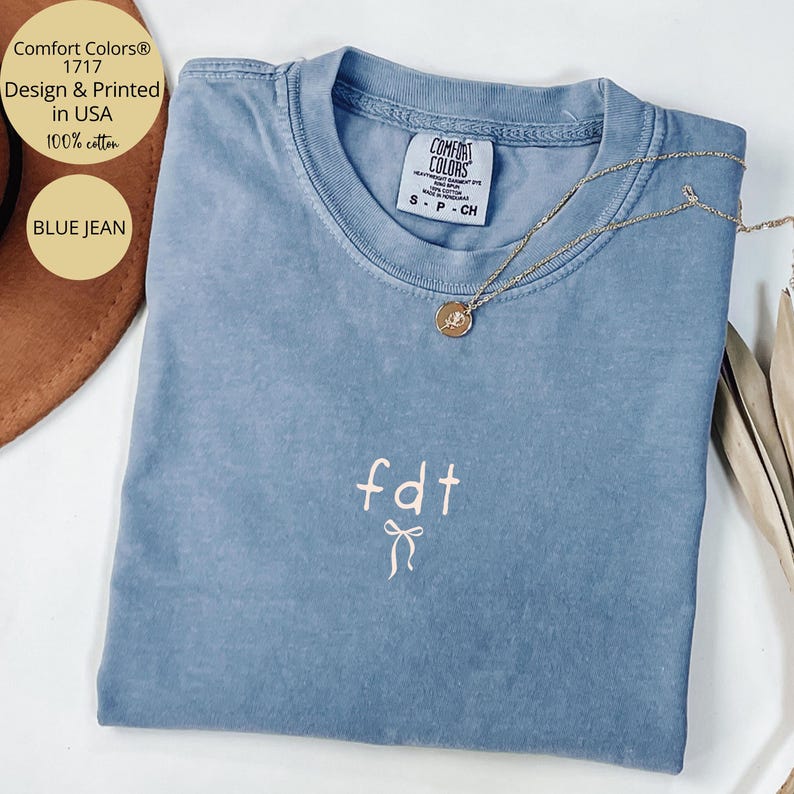Peut inclure: Un t-shirt bleu jean Comfort Colors avec le texte "fdt" et un motif de n&oelig;ud. Le t-shirt a un col rond et des manches courtes. Un collier en or avec un pendentif est drap&eacute; sur le t-shirt. Le t-shirt est en 100% coton.