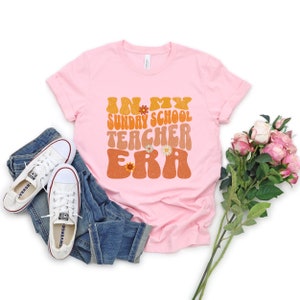 May include: Pink t-shirt with the text "In My Sunday School Teacher Era" in a retro font with flowers. The shirt is on a white background with jeans and flowers.