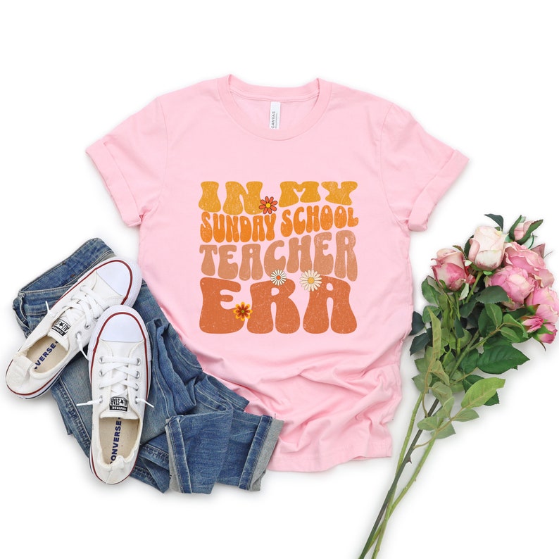May include: Pink t-shirt with the text "In My Sunday School Teacher Era" in a retro font with flowers. The shirt is on a white background with jeans and flowers.