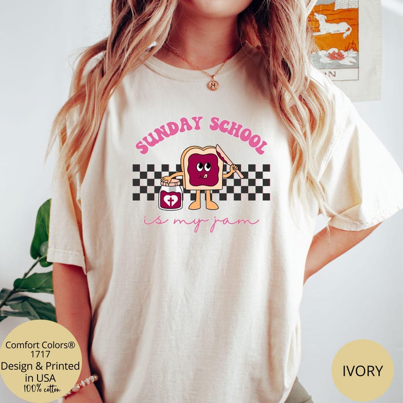 May include: A white t-shirt with the text "SUNDAY SCHOOL is my jam" in pink lettering. A cartoon illustration of a slice of bread with a face and a jar of jam with a heart on it is in the center of the shirt. The shirt is on a model and the background is a light brown color.