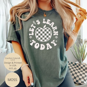 May include: Moss green t-shirt with the text "LET'S LEARN TODAY" surrounding a checkered apple graphic. The shirt is made of 100% cotton and is printed in the USA. The shirt is a Comfort Colors 1717.