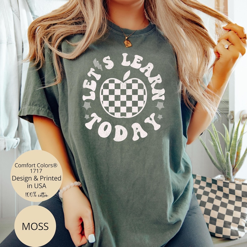 May include: Moss green t-shirt with the text "LET'S LEARN TODAY" surrounding a checkered apple graphic. The shirt is made of 100% cotton and is printed in the USA. The shirt is a Comfort Colors 1717.