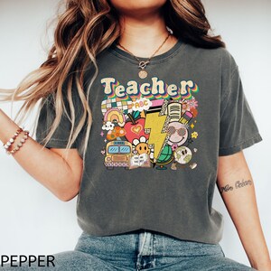 May include: A gray t-shirt with a colorful graphic design that says "Teacher" with a rainbow, school bus, apple, lightning bolt, and other school-themed imagery.