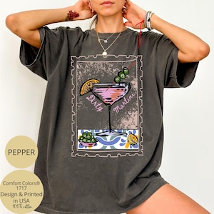 K&ouml;nnte beinhalten: Dunkelgraues T-Shirt mit einem "Dirty Martini"-Motiv im Briefmarken-Design. Die Illustration zeigt einen rosa Cocktail mit Oliven und einer Zitronenscheibe. Das T-Shirt tr&auml;gt auch den Text "Comfort Colors" und "Design & Printed in USA".