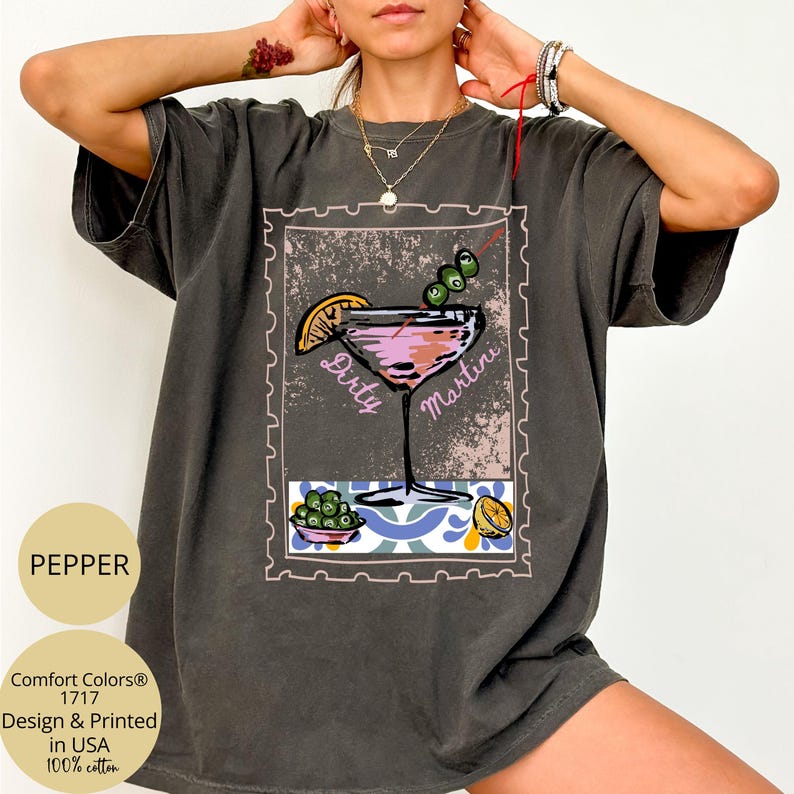 K&ouml;nnte beinhalten: Dunkelgraues T-Shirt mit einem "Dirty Martini"-Motiv im Briefmarken-Design. Die Illustration zeigt einen rosa Cocktail mit Oliven und einer Zitronenscheibe. Das T-Shirt tr&auml;gt auch den Text "Comfort Colors" und "Design & Printed in USA".