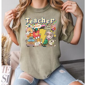 May include: A green t-shirt with the word "Teacher" in a retro font and colorful illustrations of school supplies, including a school bus, an apple, a lightning bolt, a globe, and a pencil.