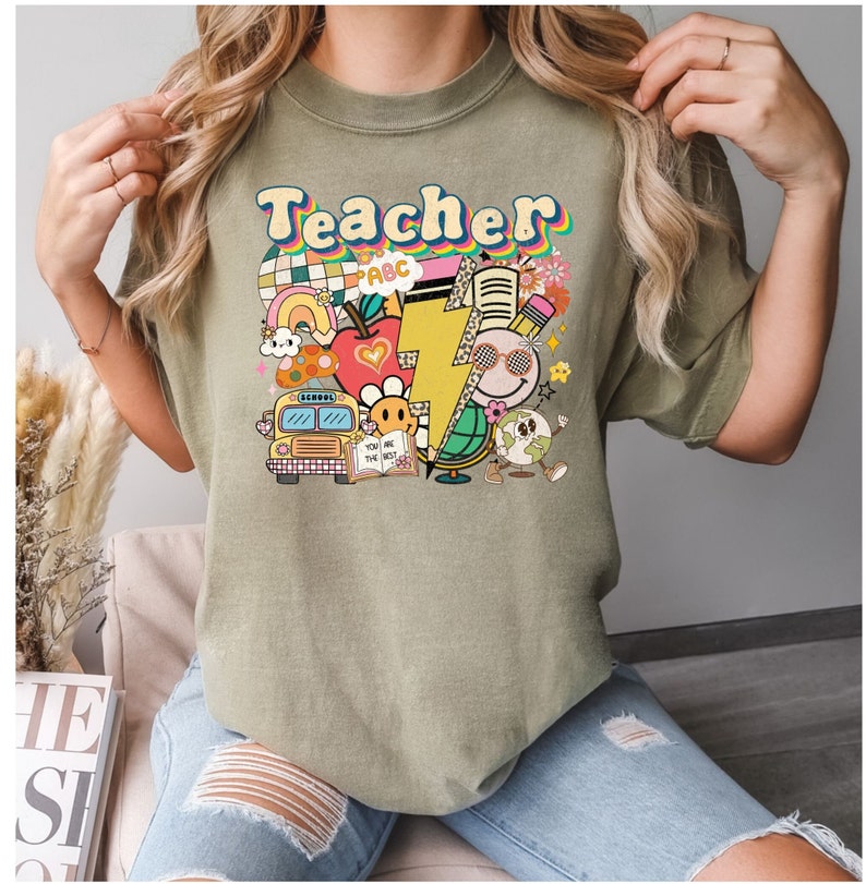 May include: A green t-shirt with the word "Teacher" in a retro font and colorful illustrations of school supplies, including a school bus, an apple, a lightning bolt, a globe, and a pencil.