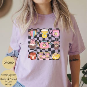 May include: A purple t-shirt with a checkered background and a design of various coffee mugs and teapots with smiley faces. The text "Comfort Colors&reg; 1717 Design & Printed in USA 100% cotton" is printed on the bottom left corner of the shirt.