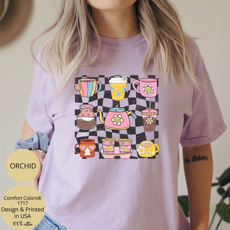 May include: A purple t-shirt with a checkered background and a design of various coffee mugs and teapots with smiley faces. The text "Comfort Colors&reg; 1717 Design & Printed in USA 100% cotton" is printed on the bottom left corner of the shirt.
