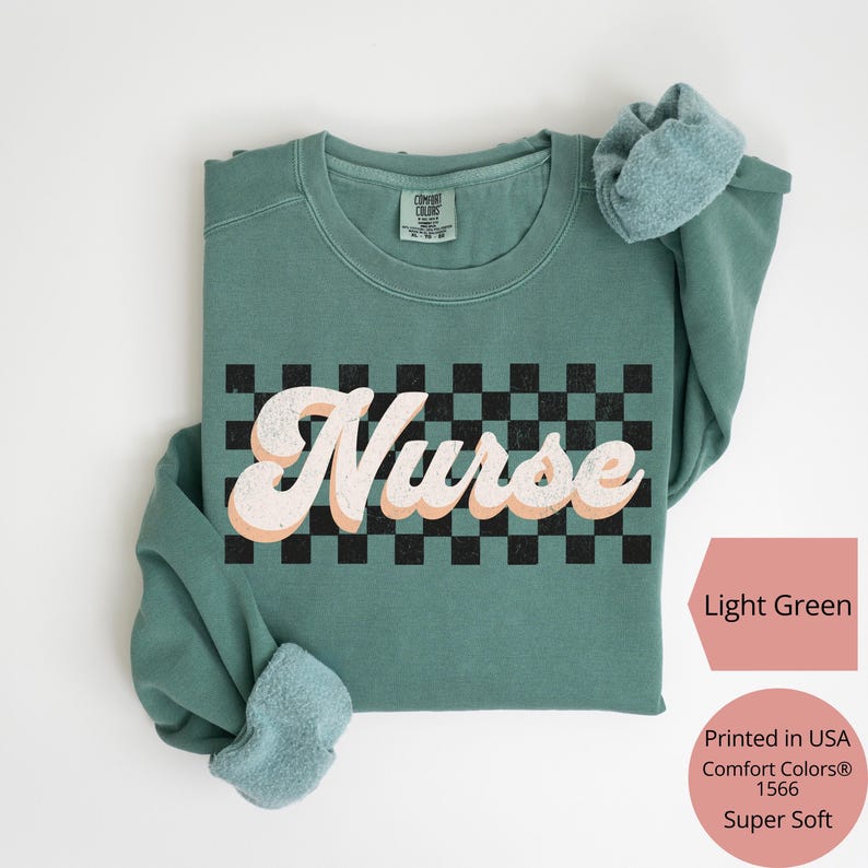 K&ouml;nnte beinhalten: Ein hellgr&uuml;nes Sweatshirt mit dem Wort "Nurse" in einem Retro-Schriftzug &uuml;ber einem schwarz-wei&szlig;en Schachbrettmuster. Das Sweatshirt ist in den USA bedruckt und besteht aus einem superweichen Material.