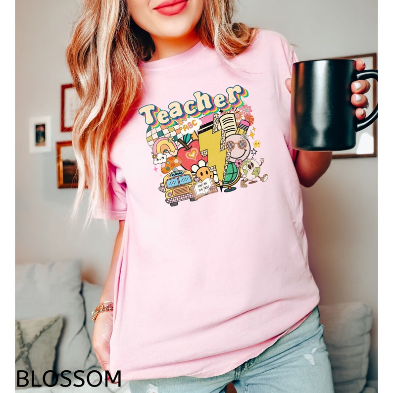 May include: Pink t-shirt with the word "Teacher" in rainbow letters and a colorful graphic of school supplies, a bus, and a smiley face.