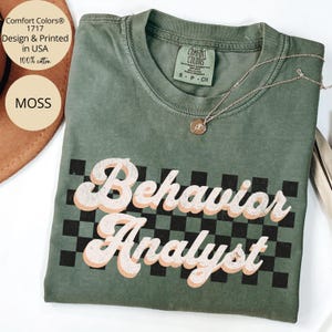 May include: A moss green short-sleeve t-shirt featuring a black and green checkered pattern background with the text Behavior Analyst printed in a distressed white and peach font. The shirt includes a Comfort Colors brand neck label.