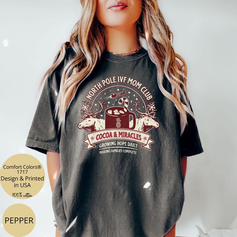 Puede incluir: Una camiseta gris oscuro con un dise&ntilde;o navide&ntilde;o que presenta una taza de chocolate caliente con malvaviscos y un bast&oacute;n de caramelo, dos renos y el texto "North Pole IVF Mom Club", "Cocoa & Miracles", "Growing Hope Daily" y "Making Families Complete".