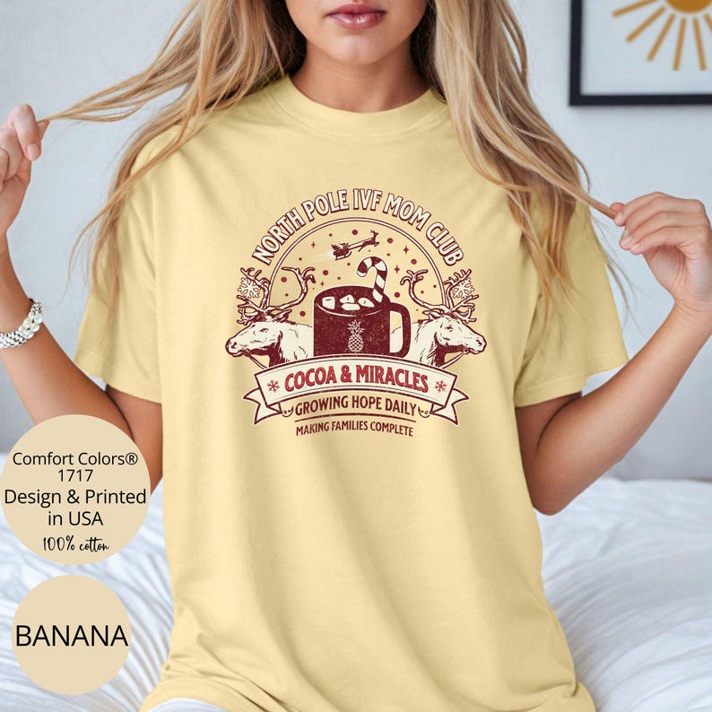 Puede incluir: Una camiseta amarilla con un dise&ntilde;o gr&aacute;fico rojo y blanco que dice "North Pole IVF Mom Club" con una taza de chocolate caliente y renos. El texto debajo del gr&aacute;fico dice "Cocoa & Miracles", "Growing Hope Daily", y "Making Families Complete".